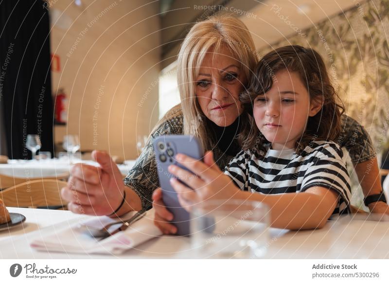 Woman and girl taking selfie on smartphone grandmother granddaughter restaurant together memory gadget take photo mobile cafe serious device table relationship