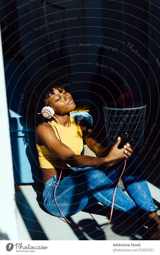 Content black woman in headphones with smartphone chilling in sunlight music listen home relax enjoy using african american song female young gadget casual rest