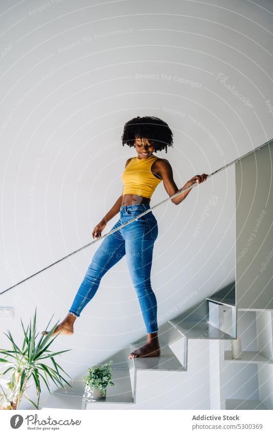Positive black woman walking down stairs afro step smile happy railing cheerful home curly hair trendy outfit casual modern female young african american