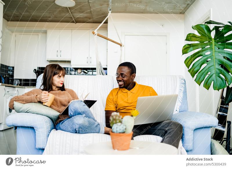 Cheerful couple with book and laptop at home read using sofa happy smile cheerful love together relationship living room multiethnic browsing diverse casual