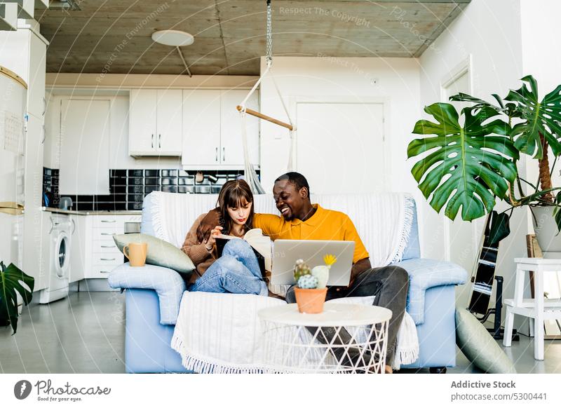 Cheerful couple with book and laptop at home read using sofa happy smile cheerful love together relationship living room multiethnic browsing diverse casual