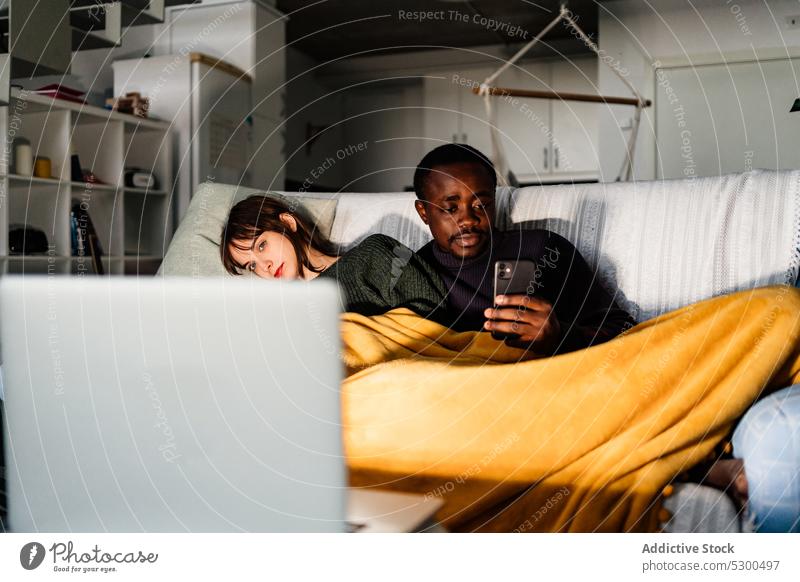 Serious black man browsing smartphone while watching movie with girlfriend couple laptop weekend spend time rest using sofa home together boyfriend relax gadget