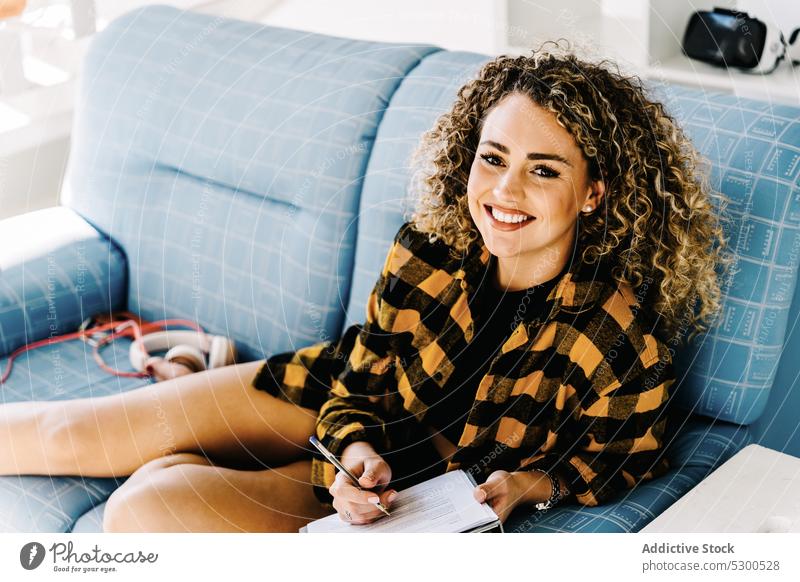 Smiling woman writing notes in clipboard take note write at home sofa weekend imagination thought compose smile positive female young curly hair checkered shirt