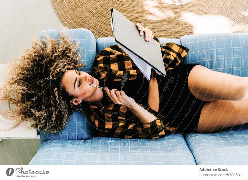 Woman writing notes in clipboard woman take note write at home sofa weekend imagination thought compose female young lying curly hair checkered shirt casual pen