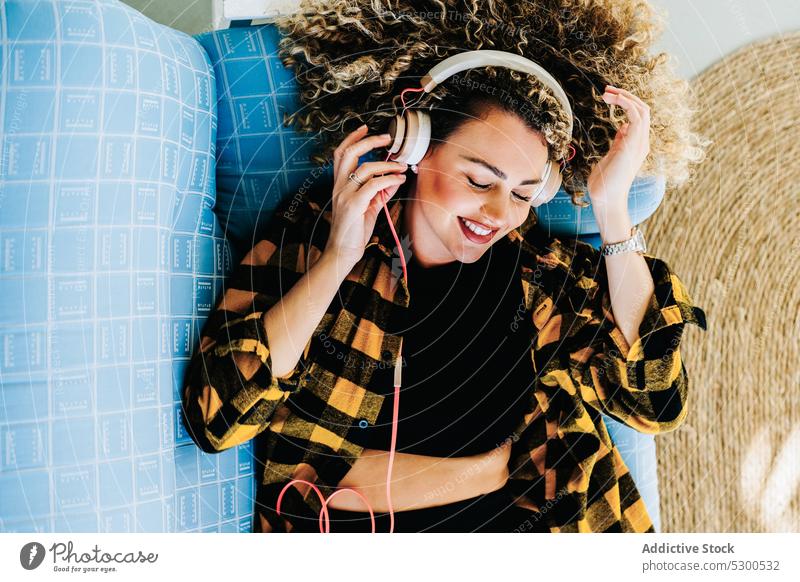 Cheerful woman in headphones lying on sofa listen music at home melody sound smile positive female young curly hair inspiration information carefree