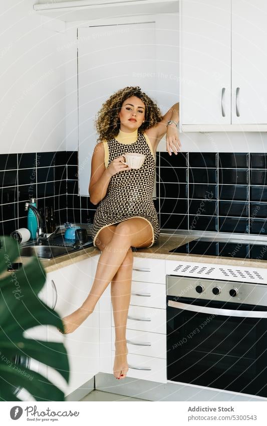 Young woman sitting on counter with cup of hot drink kitchen breakfast coffee tea morning at home female young lean on cupboard beverage dress curly hair