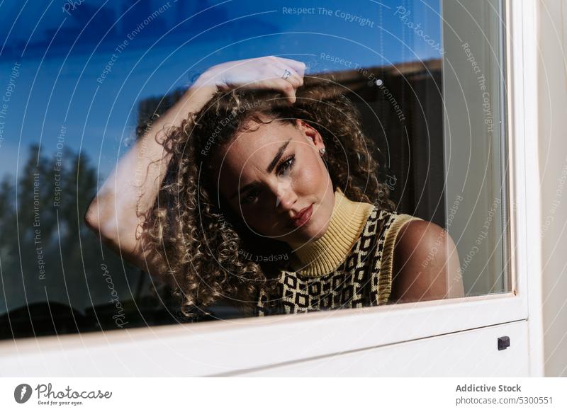 Young woman adjusting curly hair looking at camera fix windowsill free time daytime sunny touch hair reflection blue sky calm female young peaceful tranquil