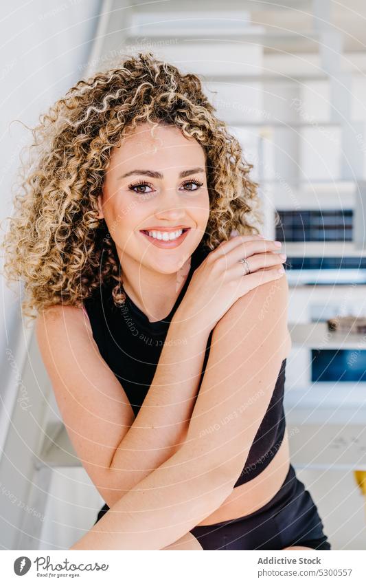 Smiling woman sitting on staircase touch shoulder glance appearance curly hair charm individuality toothy smile cheerful happy positive female young slim
