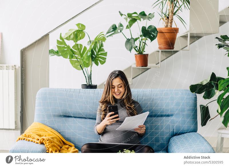Smiling pregnant woman with documents and using smartphone apartment talk conversation sofa smile female young work busy gadget communicate laptop speak