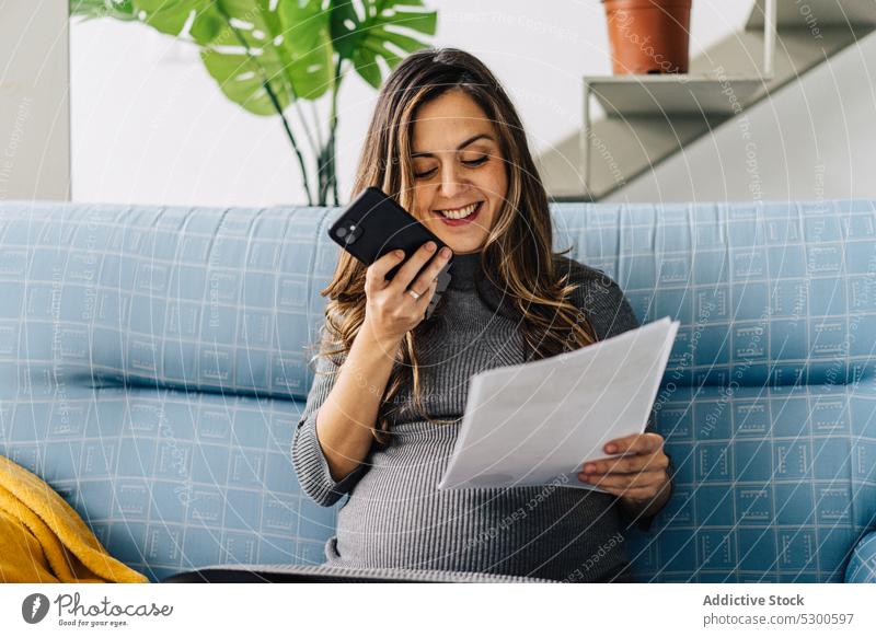 Smiling pregnant woman with documents having phone conversation using smartphone phone call apartment talk sofa smile female young work busy gadget communicate