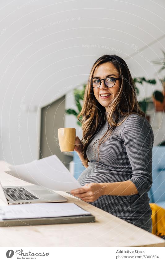 Smiling pregnant businesswoman working with documents and on laptop read using self employed remote freelance smile female young carry gadget cup at home happy