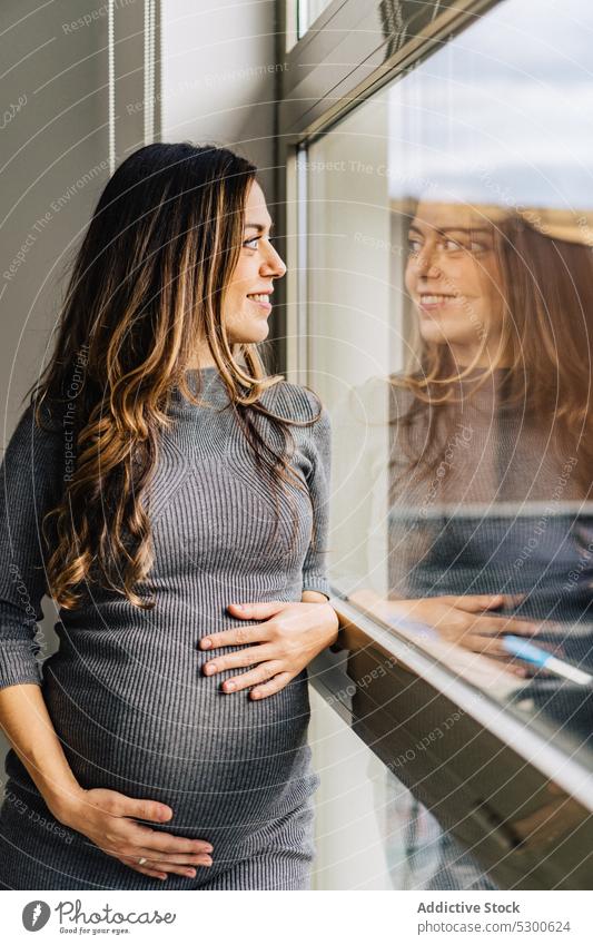 Pregnant woman standing near window caressing belly pregnant love touch belly maternity leave anticipate tranquil mother await smile female young stomach