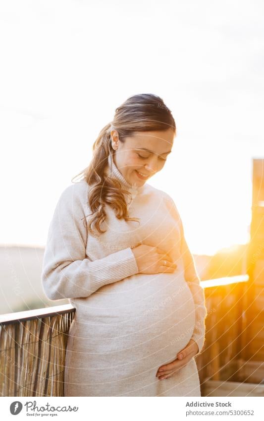 Happy pregnant woman hugging belly on terrace unborn baby tender love caress touch belly sunlight smile happy sunset female young ponytail anticipate