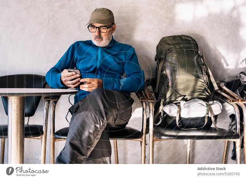 Focused senior man browsing smartphone at table message using traveler online serious trip surfing male beard mauritania backpack mobile eyeglasses casual
