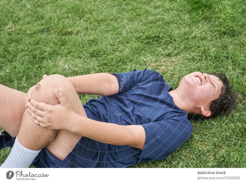 Upset boy lying on grass after injury tired rest injure suffer pain ache sportswear hurt lawn athlete eyes closed problem unhappy knee dissatisfied upset