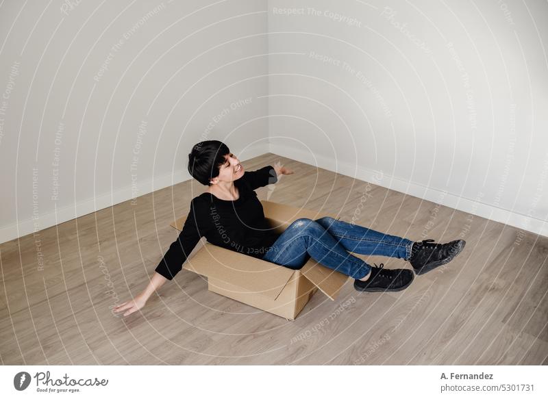 A young woman with short hair inside a cardboard box in an empty room. Concept of moving out and starting a new life. Playing with cardboard boxes, creative imagination