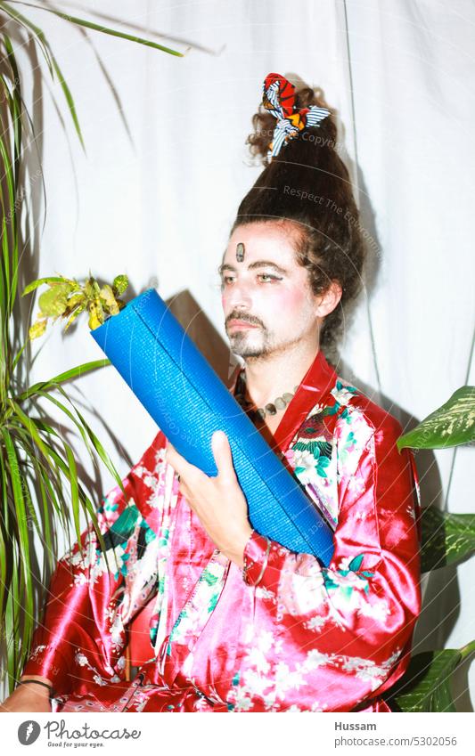 it is a photo concept about a man holding a yoga mat wearing a flowery dress and his hairy styled in a very funny way. His makeup  looks like a geisha with an ironic look.