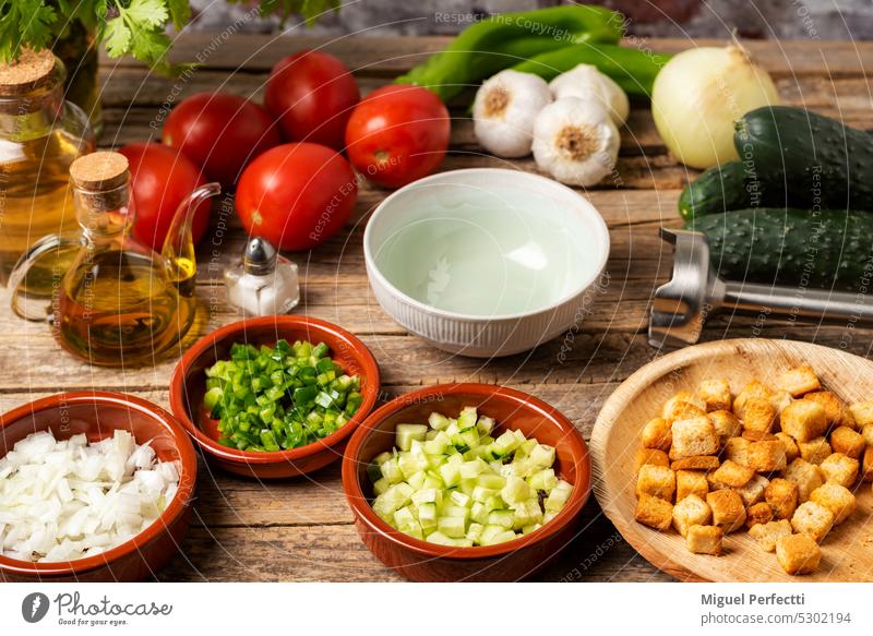 Necessary ingredients to make a gazpacho around an empty bowl, and with the chopped vegetables and toasted bread to pour it on top, on a rustic wooden table.