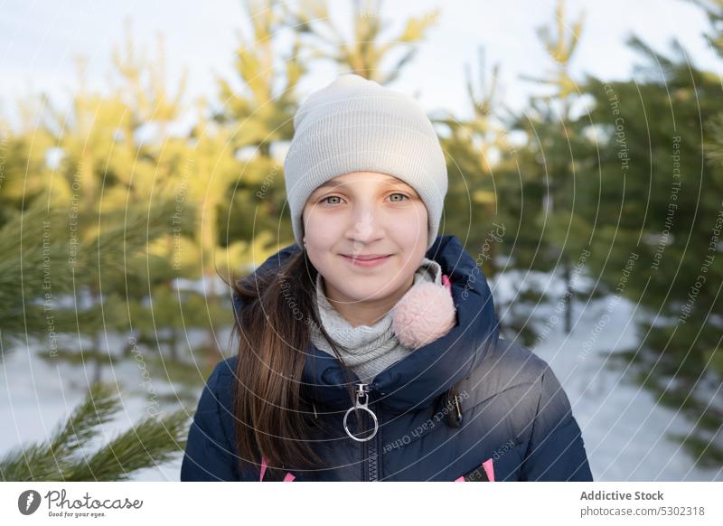 Content girl in warm clothes looking at camera in forest winter snow holiday nature tree smile cold child positive happy childhood hat preteen cheerful woods