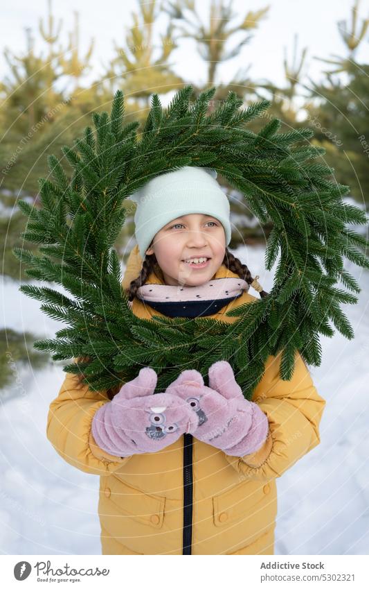 Cute preteen girl with wreath in snowy forest winter nature warm clothes smile coniferous hat cold woods season outerwear happy woodland wintertime positive