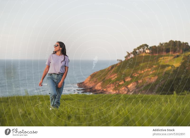 Happy woman walking on grassy hill near sea admire nature picturesque peaceful travel smile happy female casual sunglasses enjoy summer young freedom trip