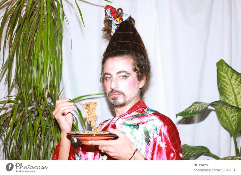 it is a photo concept about a man holding a yoga mat wearing a flowery dress and his hairy styled in a very funny way. His makeup  looks like a geisha with an ironic look. eating noodles and looking to the camera.