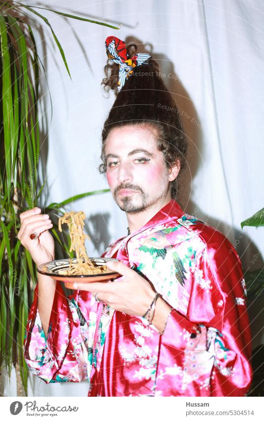 it is a photo concept about a man holding a yoga mat wearing a flowery dress and his hairy styled in a very funny way. His makeup  looks like a geisha with an ironic look and eating noodles.