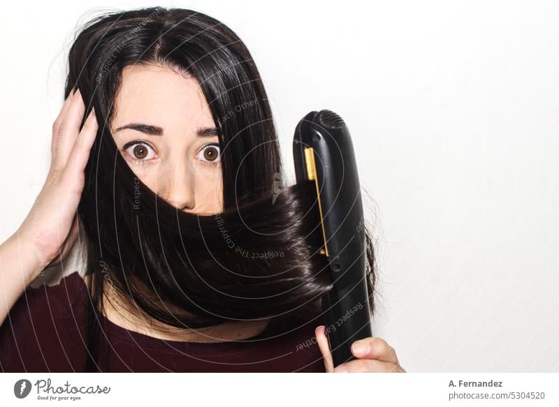 Detail of a woman with long hair and a gesture of surprise using hair straighteners on a white background. Concept of using heat to straighten hair. iron hair