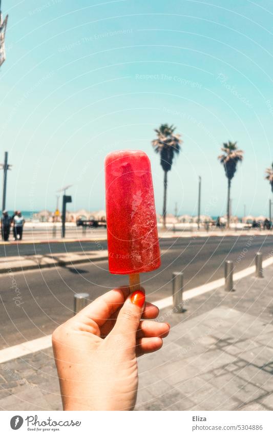 Hand holding red popsicle on a beach boardwalk on vacation. ice on a stick Ice Summer Decompose Delicious beach promenade Refreshment Summery palms Cold Frozen