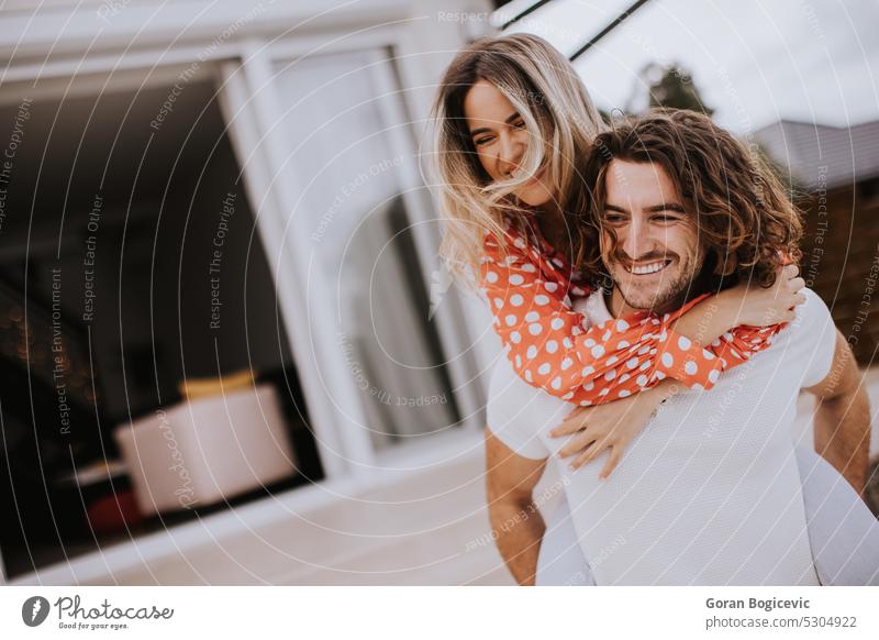 Handsome long hair man carrying the young woman on his back in front of brick house love couple happy two together people family handsome buy husband smile