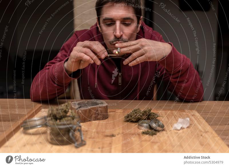 Adult man smoking cannabis at home smoke smoker marijuana light joint blunt match ganja habit male adult medical legalize nicotine weed drug wooden table addict