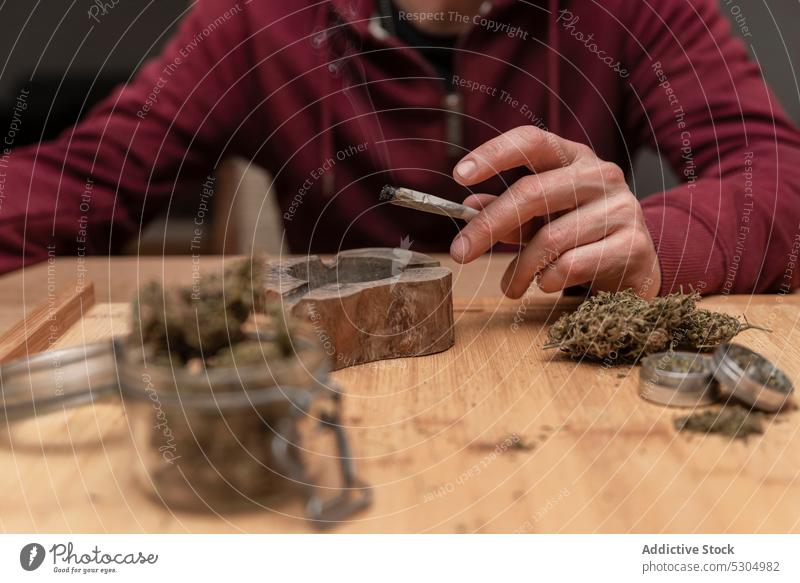 Crop man sitting at table while smoking cannabis smoke smoker blunt herb marijuana habit legalize drug male adult joint medical product narcotic enjoy free time