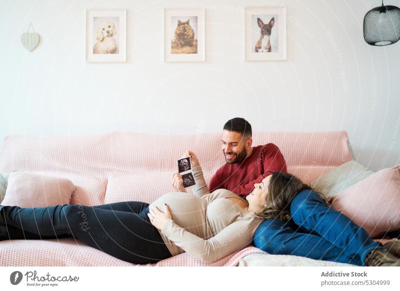 Smiling pregnant couple looking at ultrasound at home pregnancy watch love baby sofa smile together wife husband happy relationship couch anticipate affection