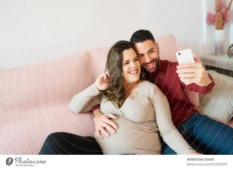 Happy pregnant couple taking selfie on cellphone smartphone together relationship happy love using bonding pregnancy cheerful sofa living room smile home belly