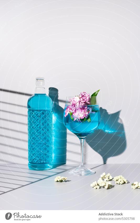 Refreshing cocktail with ice cubes near flowers and bottle drink beverage blue glass alcohol refreshment cold liquid shadow table aperitif bloom delicious serve