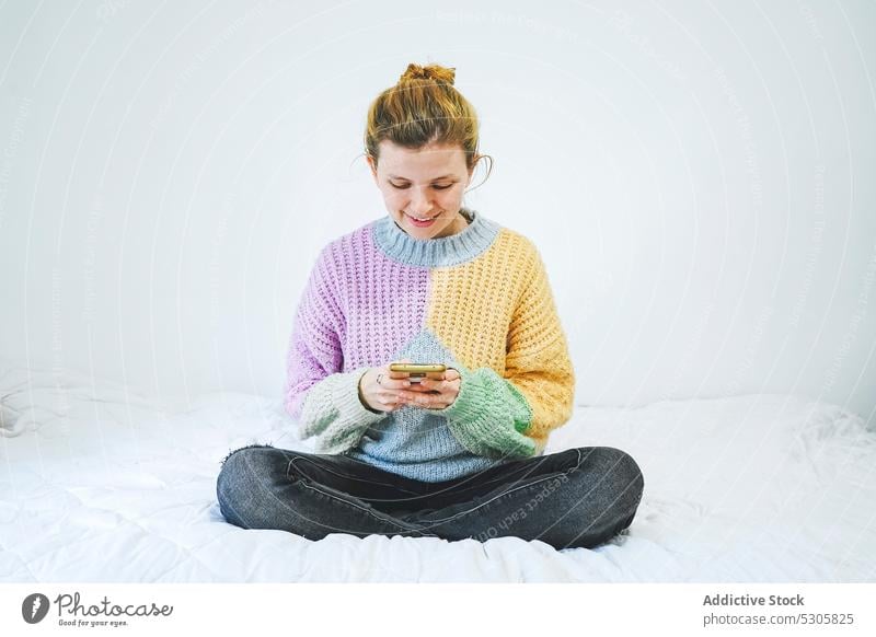 Cheerful woman browsing smartphone on bed using home smile positive happy device mobile gadget message sweater female young cheerful lotus pose padmasana