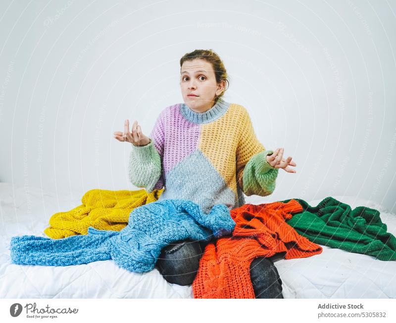 Puzzled woman with colorful sweaters looking at camera choose book bed confuse home comfort casual pensive thoughtful knitwear cloth young bedroom female
