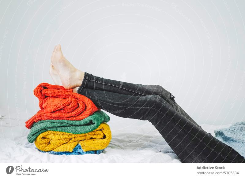 Anonymous person with legs on pile of warm sweaters colorful jeans lying cloth bed yarn knitwear home fabric soft textile comfort woolen multicolored cozy heap