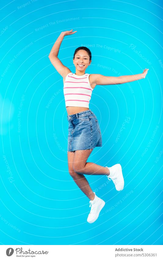 Joyful young ethnic lady jumping in blue studio woman smile arms raised excited energy cheerful model joy positive outfit happy style female mixed race brunette