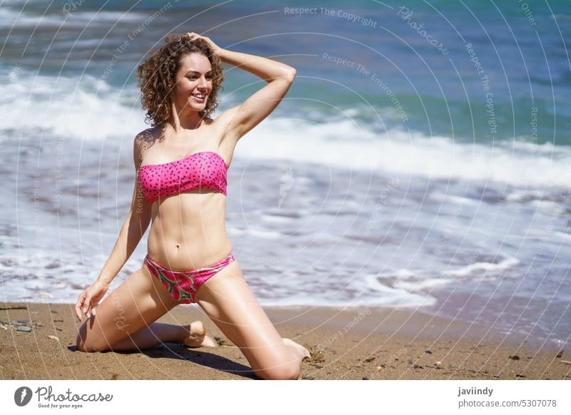 Happy woman in bikini sitting on knees on sandy beach sea touch hair smile kneel summer vacation curly hair swimwear coast female resort shore ocean happy