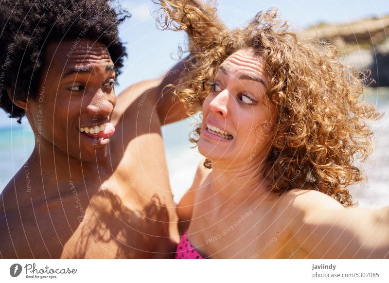 Selfie of funny diverse couple making faces on beach selfie take photo tongue out show tongue grimace make face face expression having fun touch hair cheerful