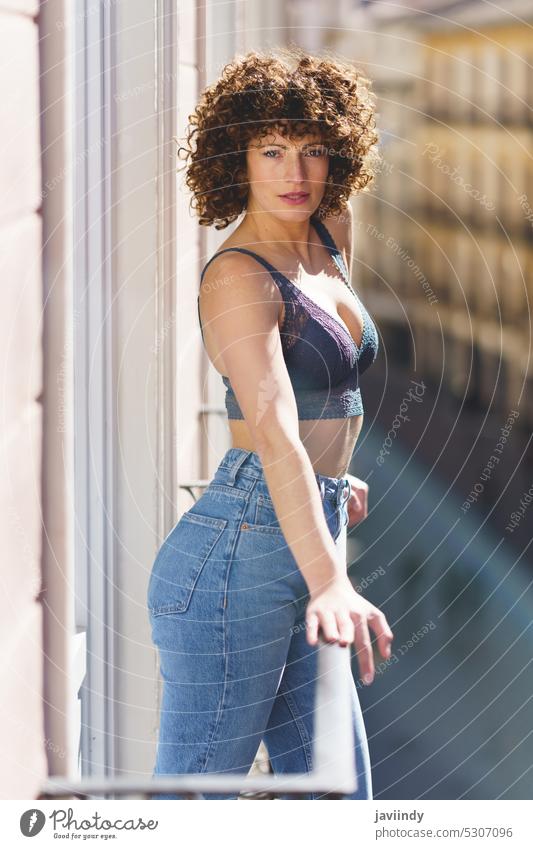 Young woman in bra and jeans looking at camera with curly hair on sunny balcony cheerful summer happy daytime terrace positive female young feminine charming