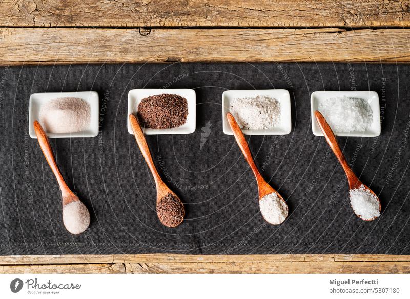 Wooden spoons next to some small plates with different types of salt, black, pink, flakes and seasoned with truffle, on a tablecloth on a rustic wooden background.Top view.