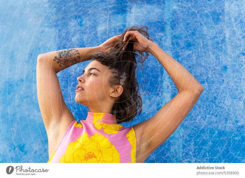 Dreamy woman touching hair against painted blue wall thoughtful street calm touch hair pensive colorful style trendy city female tattoo urban young dreamy