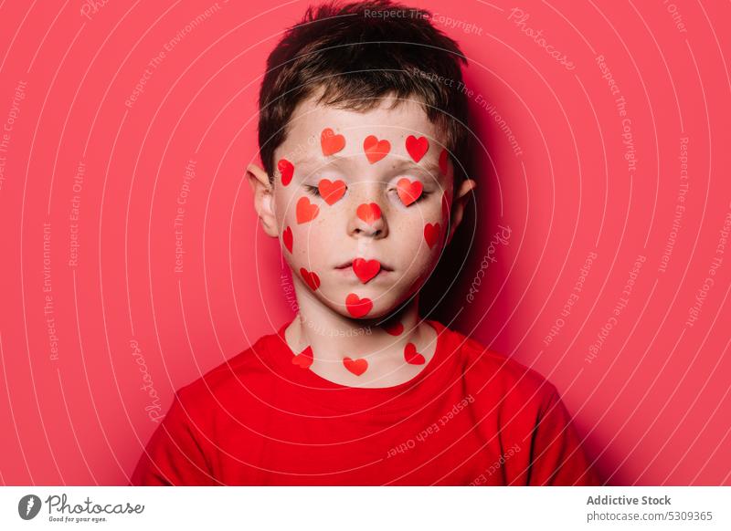 Cute boy in red casual shirt with red love sticker on face while posing for photograph kid studio cute modern adorable child childhood little colorful pensive