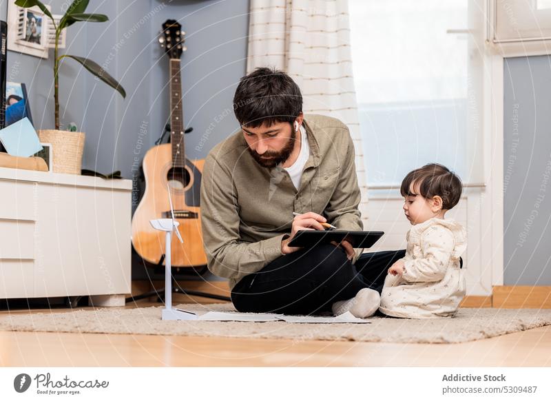Focused man examining windmill on floor near little girl tablet father daughter curious earphones project listen concentrate music child kid using home play