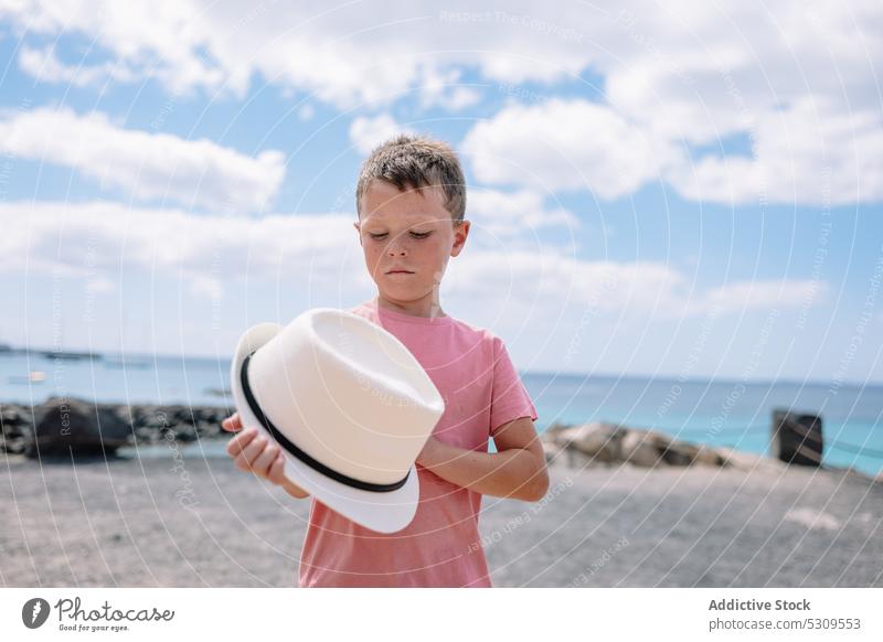Serious boy with hat standing near sea in daylight blue sky summer confident child holding hat nature ocean serious kid young childhood lifestyle beach carefree