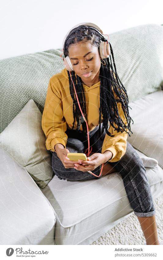 Calm black woman browsing smartphone while listening to music on sofa using home headphones surfing african american gadget female internet ethnic device young