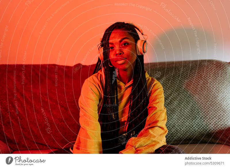 Charming African American woman listening to music in headphones on sofa neon serious home rest song meloman female braid gadget young device modern casual