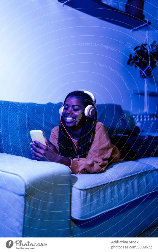 Happy black woman messaging on smartphone and listening to music on sofa headphones using lying song browsing neon happy cheerful smile gadget home device